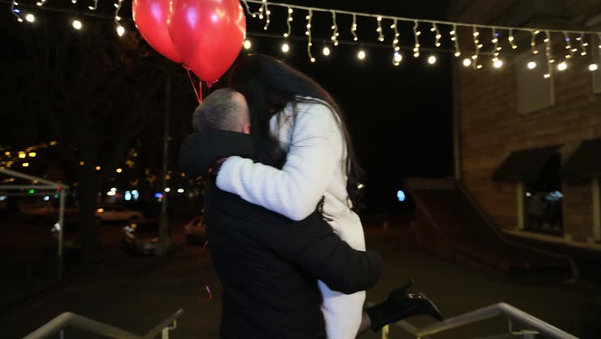 Loving couple kiss and embrace on a city street at night as the man lifts and twirling girlfriend holding red heart balloons for a joyful valentine’s day moment