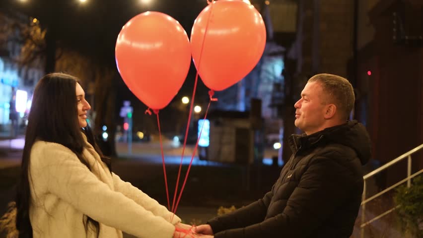 Loving couple enjoying a romantic date on a city street at night, holding red heart-shaped balloons, embracing and sharing a passionate kiss to celebrate valentine