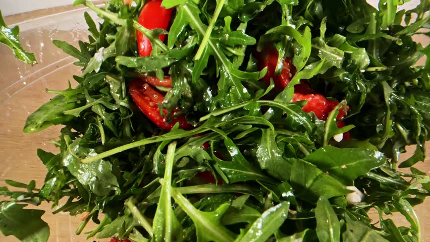 Fresh arugula salad being tossed with chopped tomatoes and dressing in a glass bowl