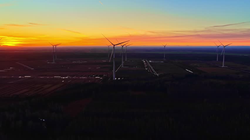 Wide drone aerial of a wind farm at sunset, turbines over farmland under an orange-to-blue sky gradient and soft haze—cinematic renewable power backdrop.