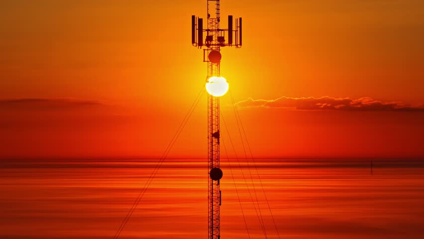 Drone ascends communication tower silhouetted against warm red sunset sun