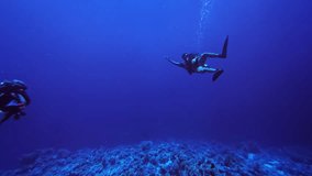 Static underwater shot of a diver floating above a deep blue reef as another diver enters the frame. Air bubbles rise toward the surface in the calm open water. - Powered by Shutterstock - Get 15% off with code: PIKWIZARD15