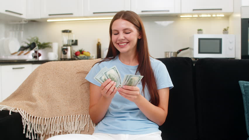 Young rich woman at home counts dollar money cash salary, earnings and smiles at total. Caucasian girl on sofa plans purchase invests feels proud about salary big bonus and dreams about new buy.