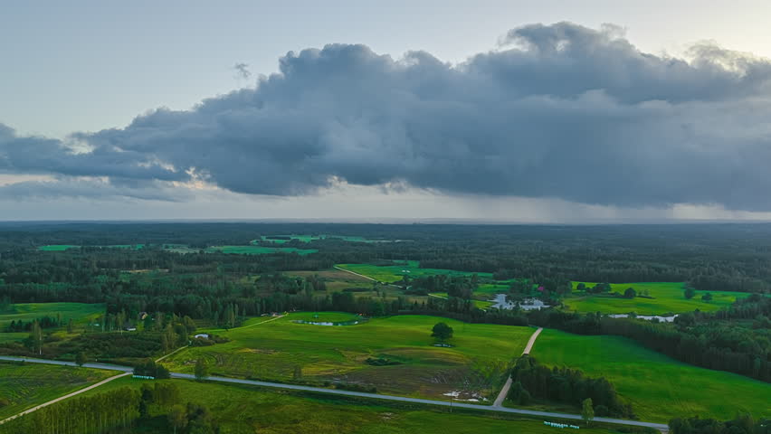 Low, fast-moving storm clouds sweep across a wide rural landscape, revealing shifting light over fields, forests, and scattered farm roads in a dramatic aerial timelapse.