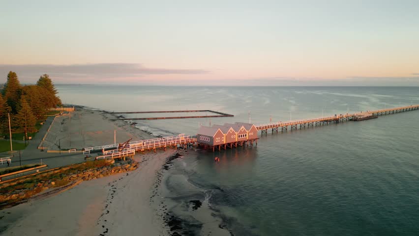 Cinematic 4K drone footage of sunrise at Busselton Jetty in Western Australia, featuring calm glassy water, a soft orange glow across the horizon, and a peaceful empty scene with no people.