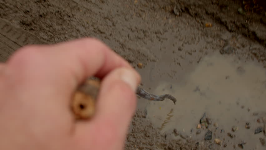 first person pokes muddy puddle with metal rod, creating ripples and splashes on clay surface, investigative motion and tactile curiosity, small tool testing water depth and soil