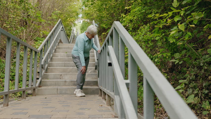 Elderly woman standing on outdoor stairs and holding her knee due to sudden pain. High quality 4k footage