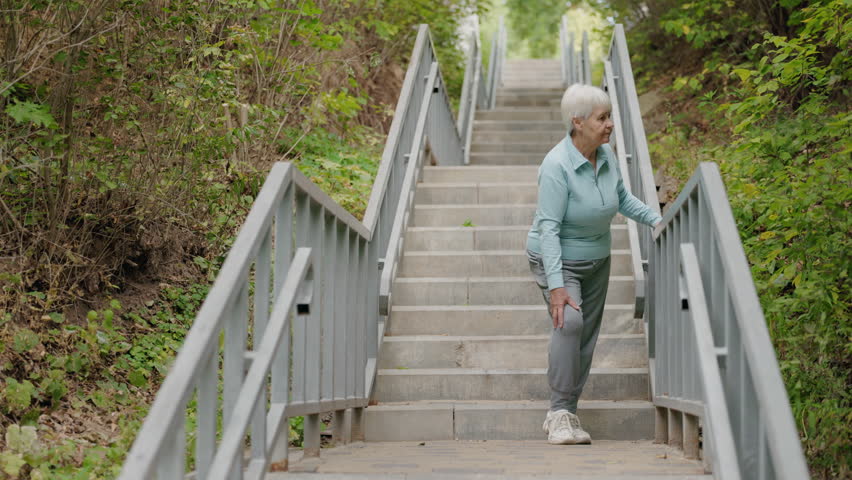 Elderly woman holding handrail while walking on outdoor stairs in a green park. High quality 4k footage