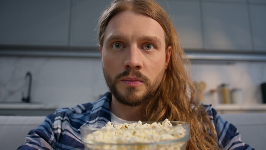 Focused Caucasian man binge eating popcorn watching TV at home close up stuffing mouth male guy overeating tasty corn snack obsessed movie television series weekend relaxation unhealthy habit indoors