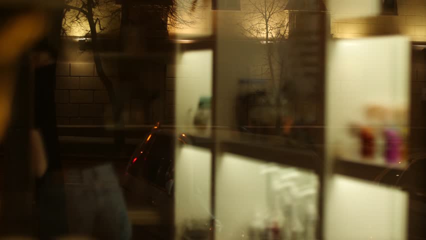 Warmly lit store interior is visible through the large nighttime window, where woman working thoughtfully. Soft reflections on glass cozy lighting create inviting, quiet urban scene. City lifestyle.