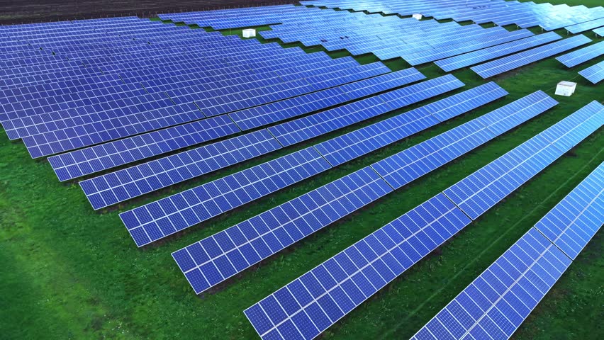 An aerial view shows numerous solar panels arrayed neatly in rows across a green field, capturing sunlight during daytime for energy generation.