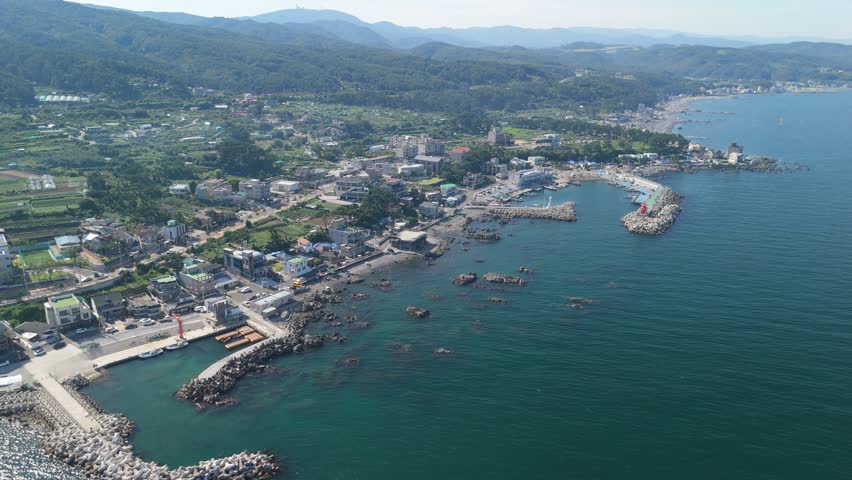 An expansive aerial view captures a picturesque coastal town nestled between the tranquil blue ocean and verdant green mountains under a bright sky. The busy harbor, adorned with boats and a distinctive red lighthouse, is protected by concrete breakwaters, while rocky shores give way to a charming beach lined with buildings. This serene landscape showcases a harmonious blend of nature and human settlement, with scattered houses and agricultural plots visible against the majestic mountainous backdrop. Ideal for travel promotions or depicting tranquil seaside communities. Concept of a peaceful coastal community.