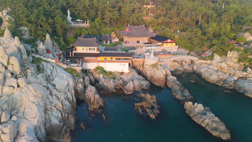 An aerial view captures the majestic Haedong Yonggungsa Temple, a renowned Buddhist sanctuary nestled along a rugged coastline in Busan, South Korea. Traditional temple buildings with ornate roofs are dramatically perched on rocky cliffs, surrounded by dense pine forests and overlooking the serene, turquoise ocean. A towering white Buddha statue (Gwanseeum-bosal) stands prominently on a hillside, while a golden Maitreya Buddha greets visitors at the temple