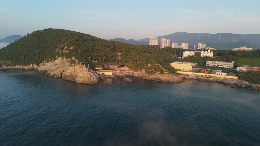 An aerial view captures the serene Haedong Yonggungsa Temple, a historic Buddhist temple nestled on a rocky coastline in Busan, South Korea. The temple complex, featuring traditional Korean architecture and a large white Buddha statue, is surrounded by lush green pine-covered hills, with the calm blue ocean water in the foreground and modern city buildings visible in the distance. Concept of spiritual tranquility and natural beauty.