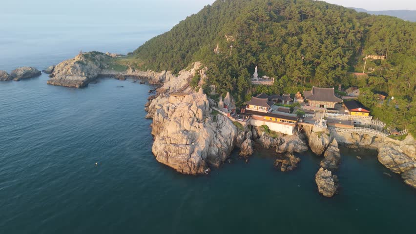 An aerial view captures the majestic Haedong Yonggungsa Temple complex nestled on a rugged, pine-covered coastline overlooking the deep blue waters of the East Sea in Busan, South Korea. Traditional Korean temple architecture with dark tiled roofs and vibrant details stands in harmony with the natural beauty, featuring a prominent white Buddha statue atop a rocky outcrop and distant city buildings on the horizon. This serene scene evokes a concept of spiritual tranquility and natural grandeur.