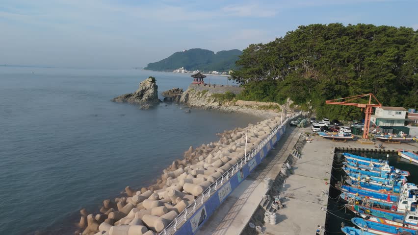 An aerial view captures a beautiful South Korean coastal landscape featuring a traditional red-roofed pavilion perched on a rocky island, lush green mountains, and a tranquil sea. The foreground showcases a robust concrete tetrapod breakwater protecting a small fishing harbor with colorful boats and an orange crane, contrasting with modern city skylines in the distant hazy background. This image evokes a sense of peaceful coexistence between nature, tradition, and modern development along the shoreline. Peaceful coastal living and urban development concept.