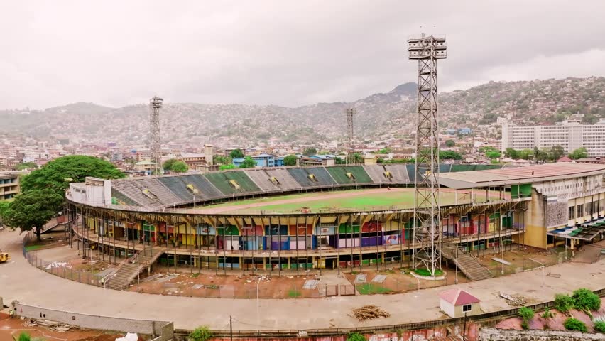 Aerial flyover revealing a dilapidated sports stadium in Freetown, Sierra Leone, showcasing abandoned architecture, overgrown grounds, urban surroundings, and decayed infrastructure, highlighting urban decay and historical sports site from above. 30 July 2025