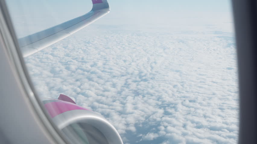 Looking out a passenger window at the airplane wing and engine soaring high above white fluffy clouds. This aerial travel scene captures the atmosphere of a commercial flight, holiday or trip.