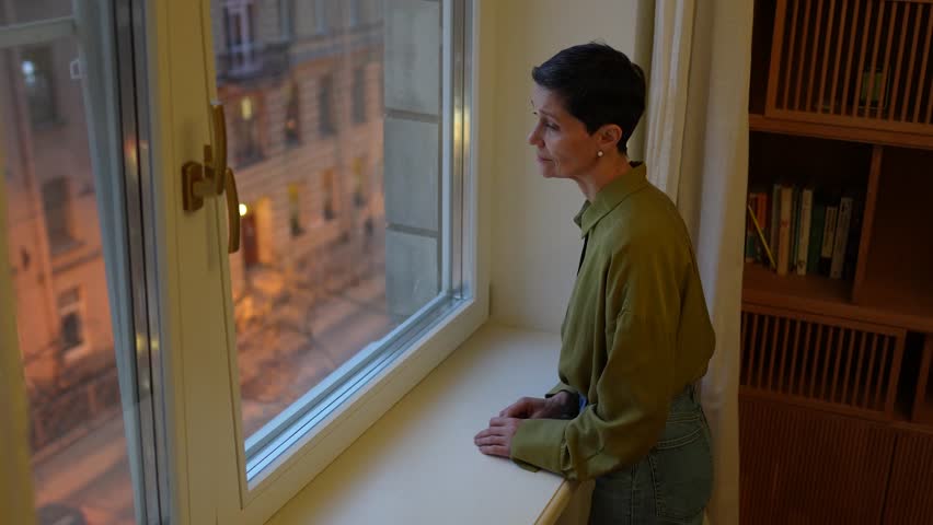 Beautiful senior woman with sad depressed expression looking outside window from dark room, mature lady dealing with elder life crisis and loneliness, coming with apathy and problems in relationships.