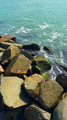 Bright sunlight dances on the turquoise water as gentle waves meet the rugged rocks covered in soft green moss, creating a tranquil seaside scene.