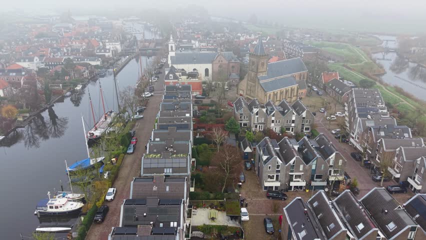 Aerial view of foggy Dutch canal town gabled brick homes, docked boats, and tall church tower form a tranquil, symmetrical waterfront scene softened by mist.
