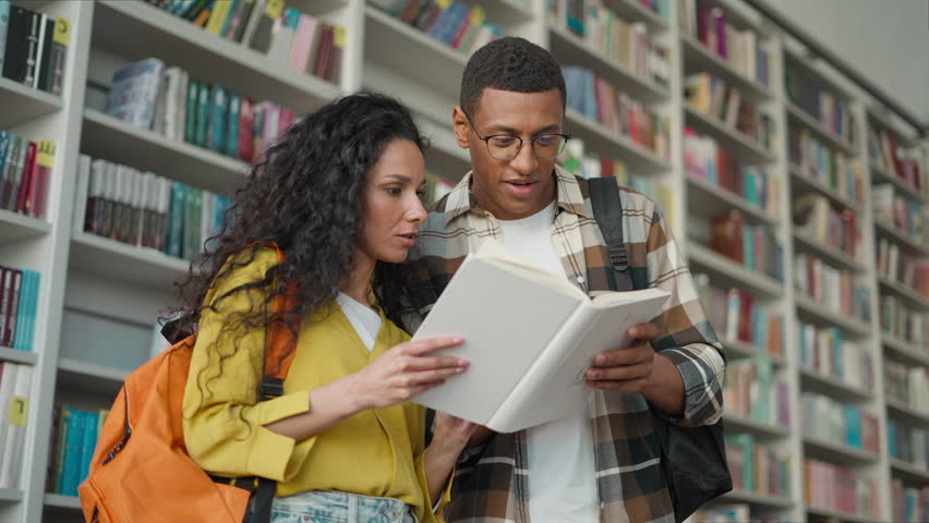 Multiethnic man woman students excited reading book together in library smiling African American male Hispanic female classmates read interesting story bookshelves education teamwork study literature