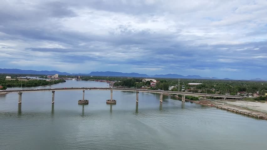 Aerial view of a long concrete bridge crossing a calm river, surrounded by lush green landscape and distant mountains under a dramatic cloudy sky. Peaceful riverside scenery with urban.