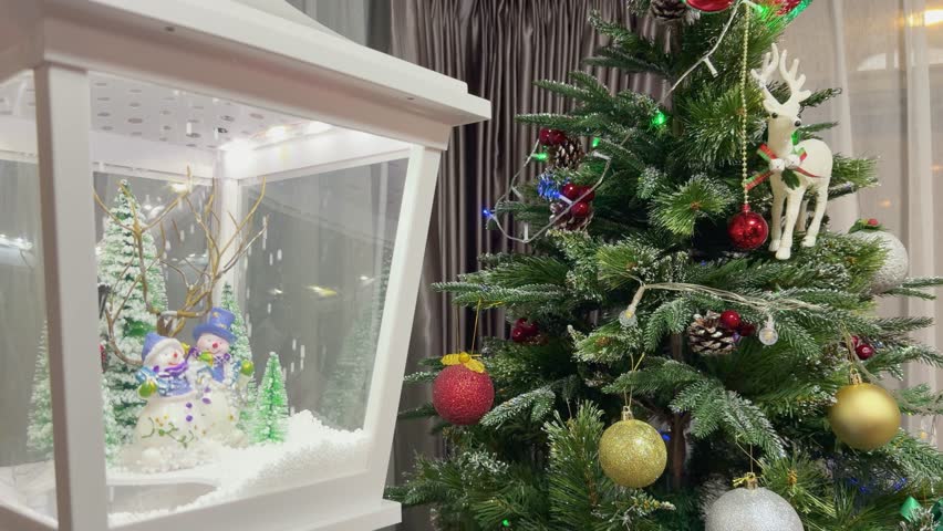 A decorated Christmas tree stands beside a glowing white lantern containing a winter snowman scene