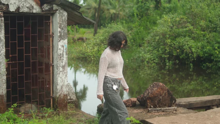 Young woman standing near a rural house by the water, looking around as if searching for something in a quiet natural setting.