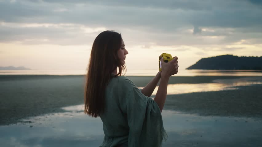 Peaceful moment of creative focus. Young woman photographer taking pictures of beautiful sunset at beach using compact digital camera, enjoying peaceful atmosphere and capturing stunning colors of sky
