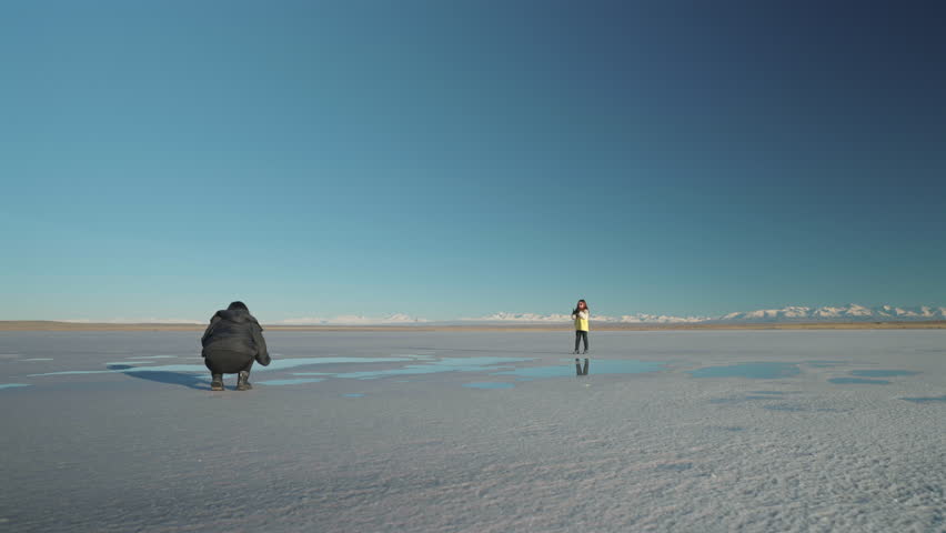 Slow motion landscape shot capturing mother or parent crouching to photograph their child jumping over shallow water on a vast, white salar, with the snowy Andes mountain range on the horizon.