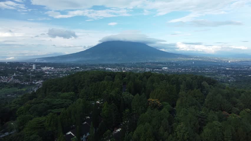 Aerial view of a volcanic mountain covered by clouds with urban area and green forest below in West Java, Indonesia. Natural landscape showing harmony between city and nature.