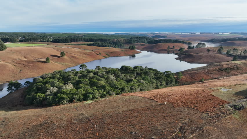 High-altitude aerial sideways reveal of a concrete dam and a full reservoir lake. Features water reflections of the blue sky within a vast rural landscape. Ideal for ESG and utility themes.