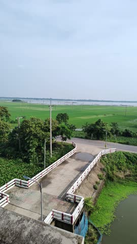 Scenic semi-rural landscape with green fields, winding road, parked car, walking person, bridge with white-red barriers, dense trees, and distant river under bright sky.