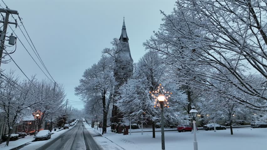 Quiet American street in winter with snow-covered trees, glowing lanterns and festive snowflake lights. Peaceful seasonal atmosphere around historic buildings at dusk. Pov drone shot.