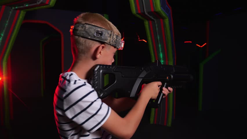 Close-up of an active boy playing laser tag with friends, he holds a laser weapon and takes aim. A game arena with bright stripes on the wall. An active play area for children.