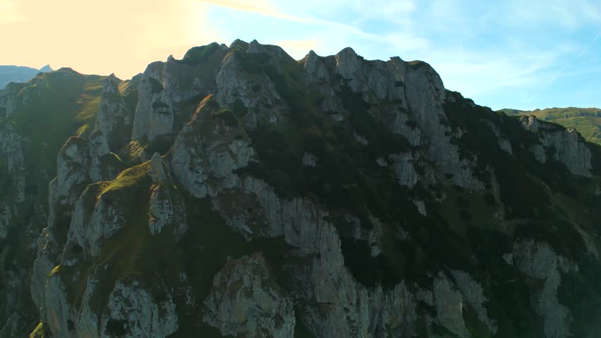 4K aerial drone shot performing an orbit maneuver around massive limestone rock formations in the Malaiesti Glacial Valley, Bucegi Mountains, Romania. This cinematic parallax sequence highlights the rugged textures and impressive geology of the Carpathian alpine landscape, set against a backdrop of distant peaks and blue sky.