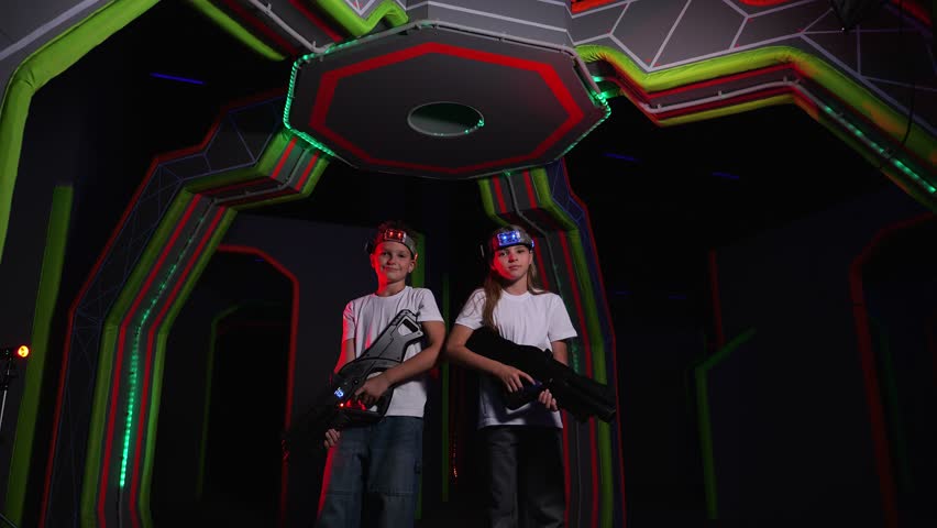 A group of cheerful teenagers with laser guns are having fun in a dark laser tag arena, a boy and a girl are standing with guns in their hands against a wall with neon stripes.
