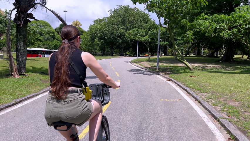 A woman cycles along a sunlit paved path in the Aterro do Flamengo, Rio de Janeiro. The shot features a wide-angle view of the park's iconic tropical trees, green lawns, and blue sky.