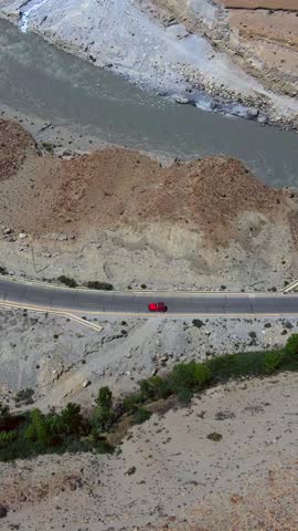 Aerial view of a red car driving on a road through a rugged landscape, with a silver river flowing nearby, Passu, Gilgit Baltistan, Pakistan.