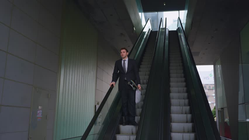 Slow motion parallax shot of professional businessman in dark suit carrying skateboard and luggage while descending on escalator in modern glass building during daily city commute