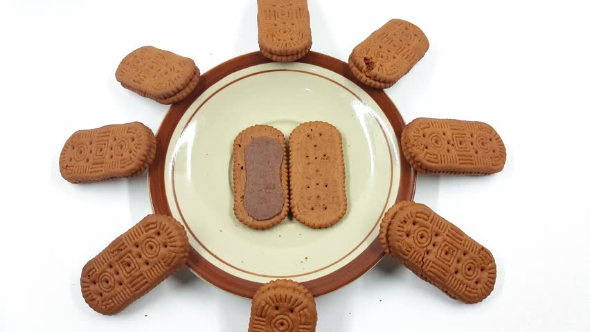 Stacked and scattered chocolate sandwich biscuits surround a small ceramic plate displaying two halves of a broken cookie.
