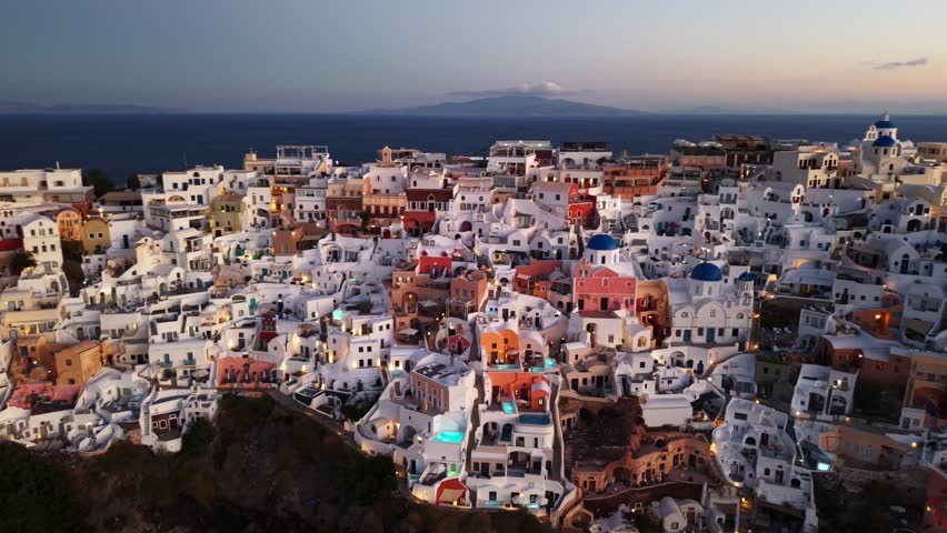 Aerial view of the dazzling white buildings of Oia cascade down the cliffs, lit by the warm glow of a setting sun, Oia, Thira, Greece.