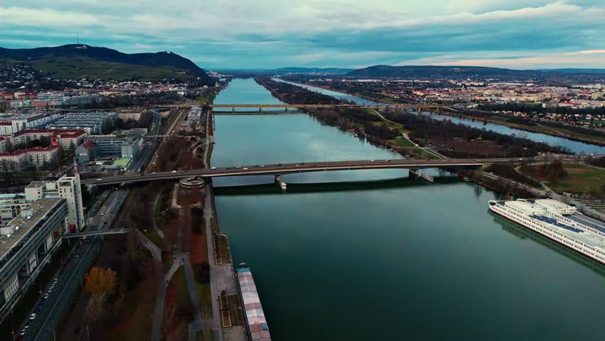Aerial landscape drone shot panning right over the Danube River in Vienna at winter sunset, showing bridges spanning the river, boats on the water, surrounding city buildings, and clouded skies over