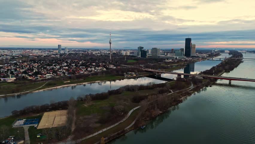 Aerial landscape drone shot panning left over the Danube River in Vienna, showing the river, surrounding cityscape, bridges, buildings along the banks, and muted winter light with cloudy skies over