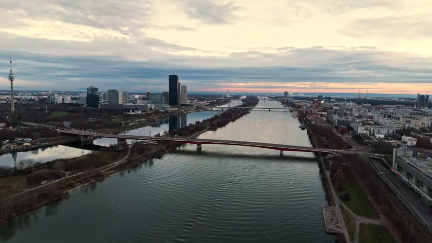 Aerial landscape drone shot over the Danube River in Vienna at winter sunset, showing the river, bridges, surrounding city buildings, boats on the water, and clouded skies overhead.