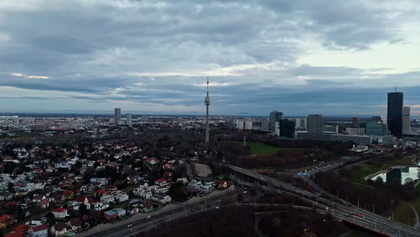 Aerial landscape drone shot panning right toward the Danube Tower in Vienna, showing the tower, surrounding city buildings, riverbanks, and muted winter light with cloudy skies overhead.