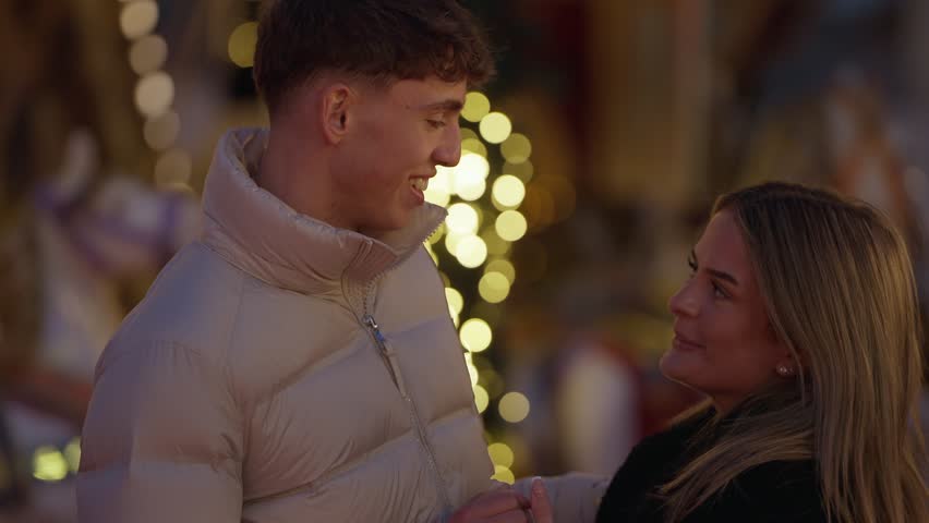 A young couple in love holds hands and looks at each other in Paris France at night Bokeh lights create a romantic atmosphere Medium handheld shot.