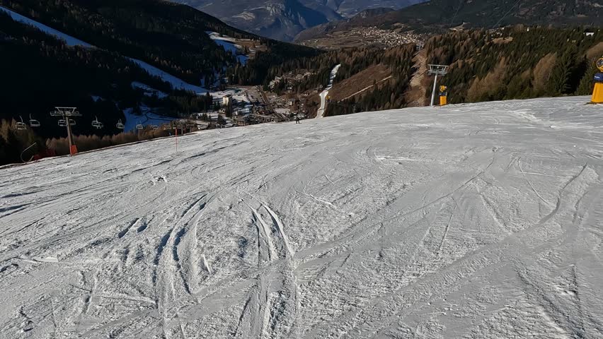 First person view standing on a ski slope looking down towards a valley village surrounded by majestic snowy mountain ranges in Italy Folgaria