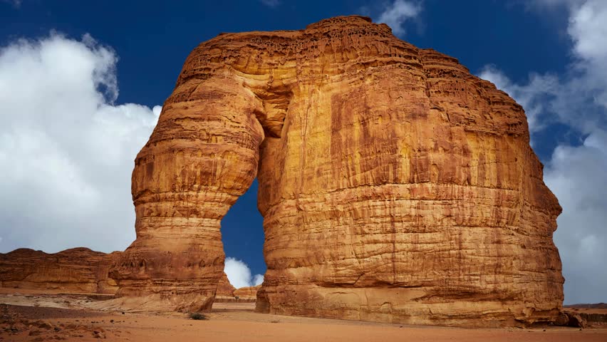 Elephant Rock Geological Formation in AlUla Desert 

iconic landmark, geological wonder, natural formation, desert landscape, rock structure, tourism, destination, 
A striking shot of "Jabal Al-Fil" (Elephant Rock) in AlUla, Saudi Arabia. This massive natural red sandstone monolith is shaped like an elephant with a trunk reaching the ground. The video captures the golden hour tones of the desert sand and rock under a scattered cloudy sky.